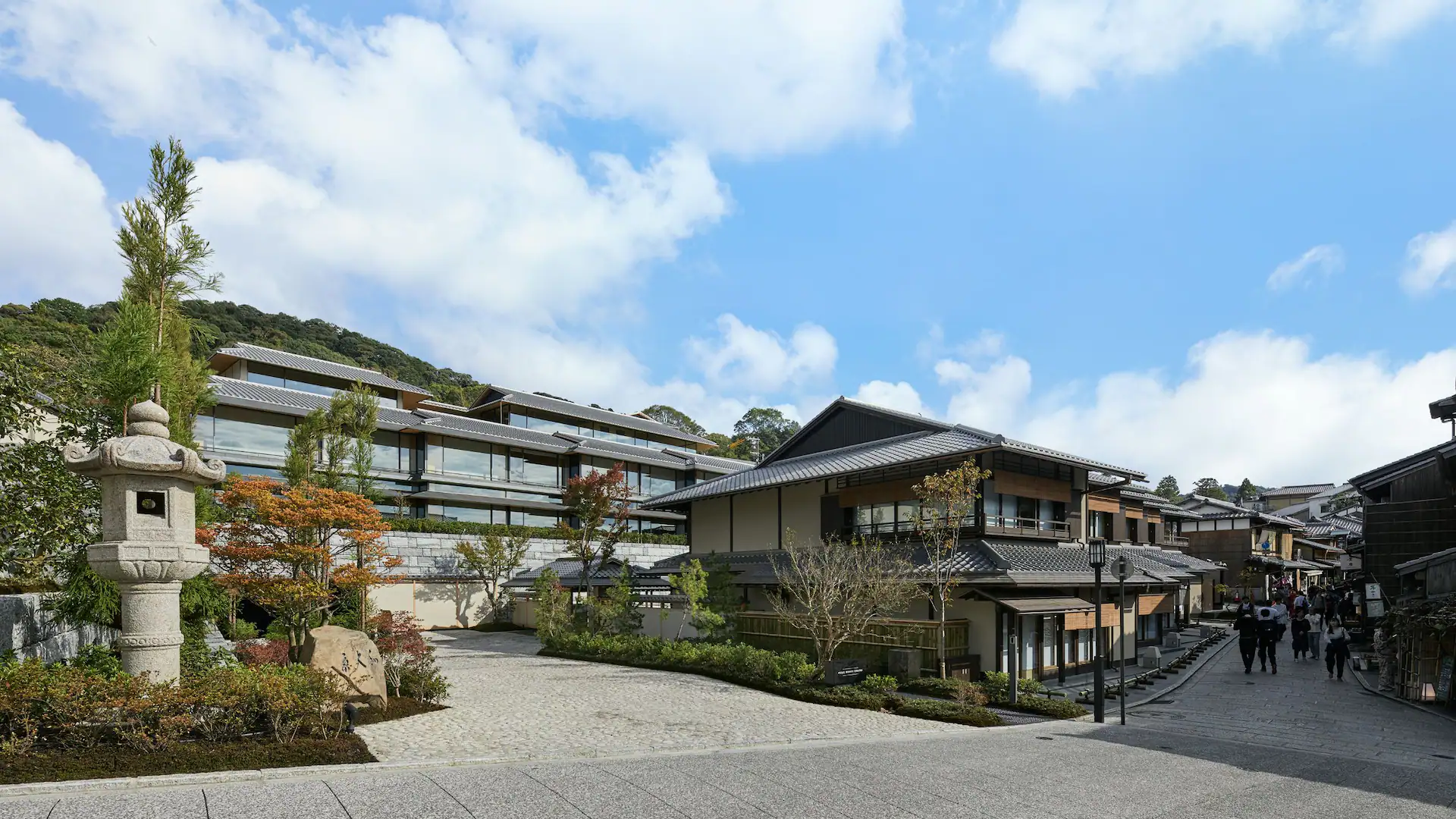 Traditional Japanese street with stone lantern, wooden houses, and vibrant trees. People stroll under a clear blue sky. Peaceful atmosphere.
