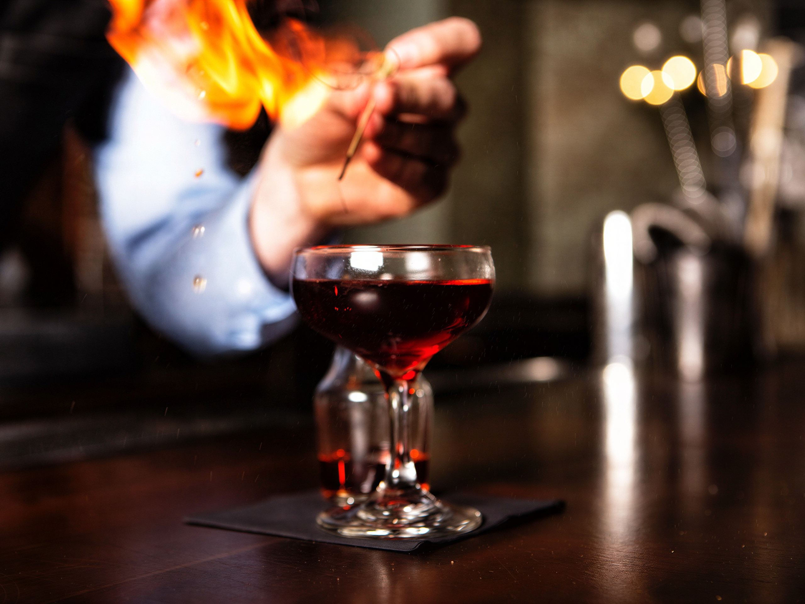 A hand ignites a drink above a glass with a flame in a dim bar setting, creating a warm and dramatic effect. No visible text.
