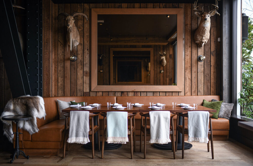 Cozy restaurant with wooden walls, leather bench, and a set table. Animal mounts and large mirror enhance a rustic, inviting ambiance.