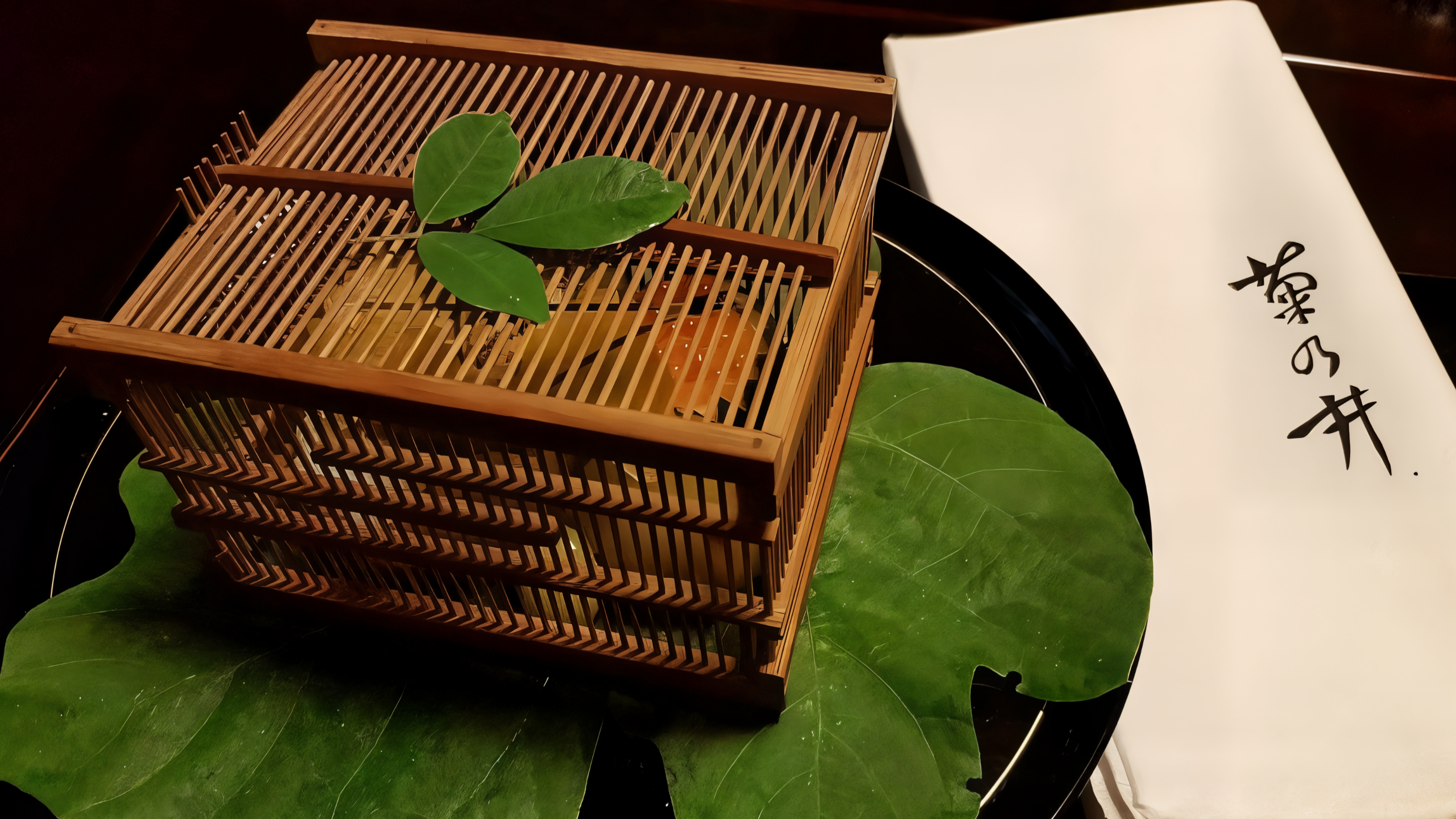 Wooden lattice box with green leaves on top, placed on large green leaves, next to a white napkin with Japanese text. Elegantly presented.