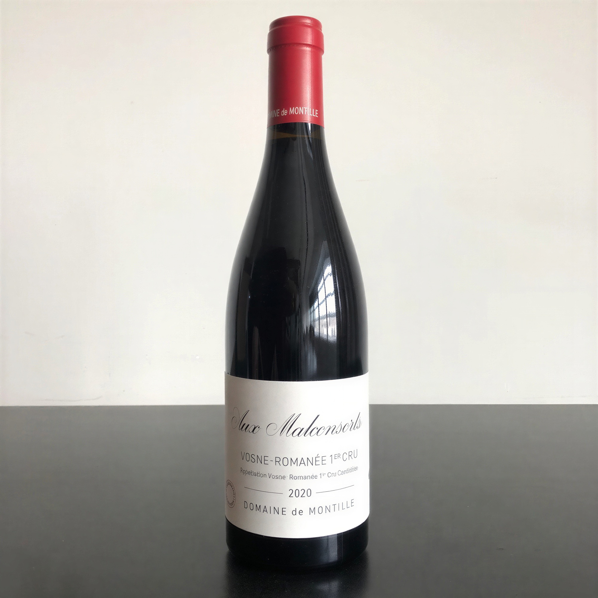 A bottle of Domaine de Montille Aux Malconsorts 2020 wine, with a red cap, stands on a dark surface against a plain background.