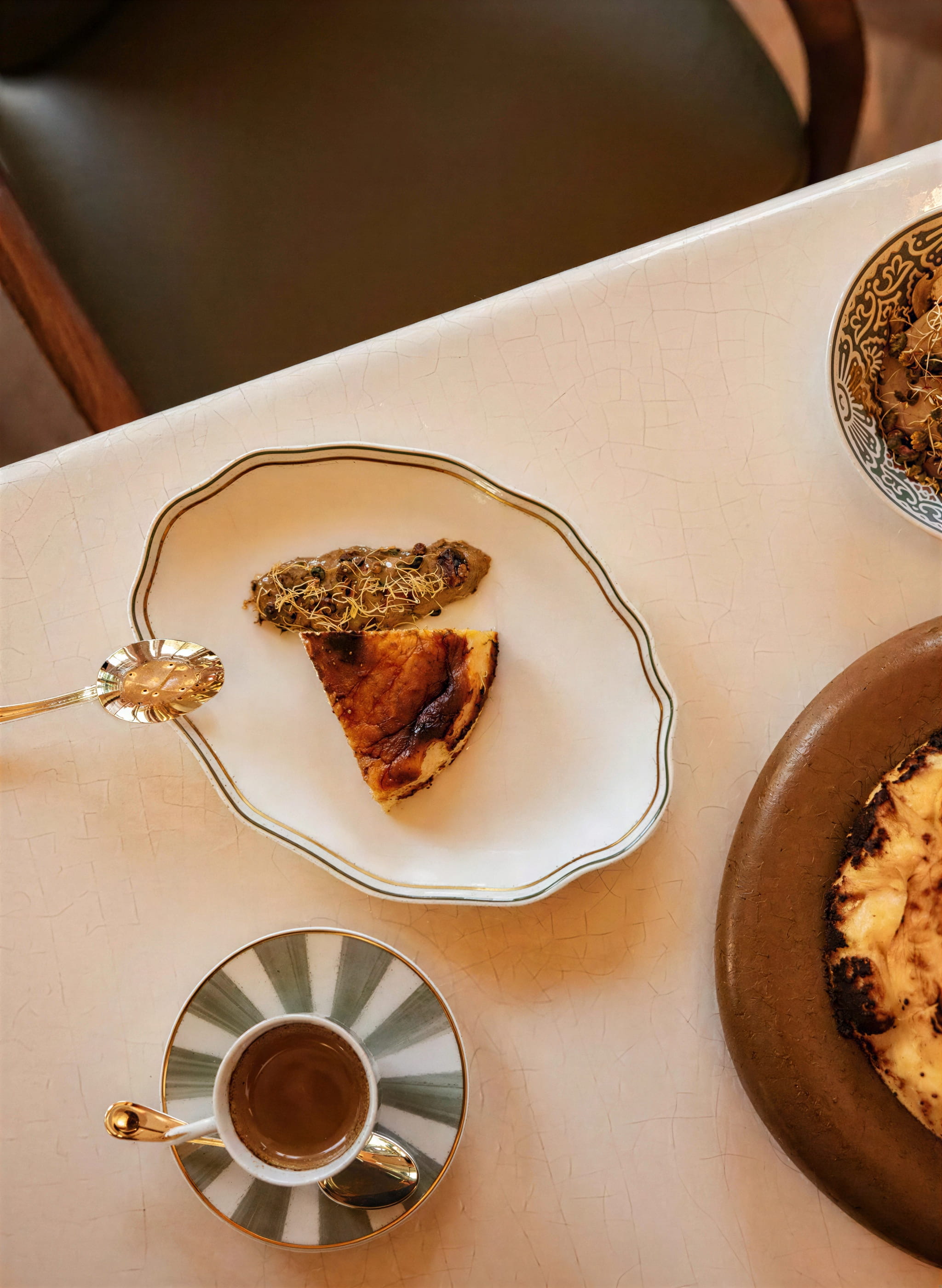 Slice of pistachio honey cheesecake from Serra at The Chancery Rosewood and topping on a decorative plate, next to a green-striped cup of espresso on a white table. Cozy, inviting atmosphere.
