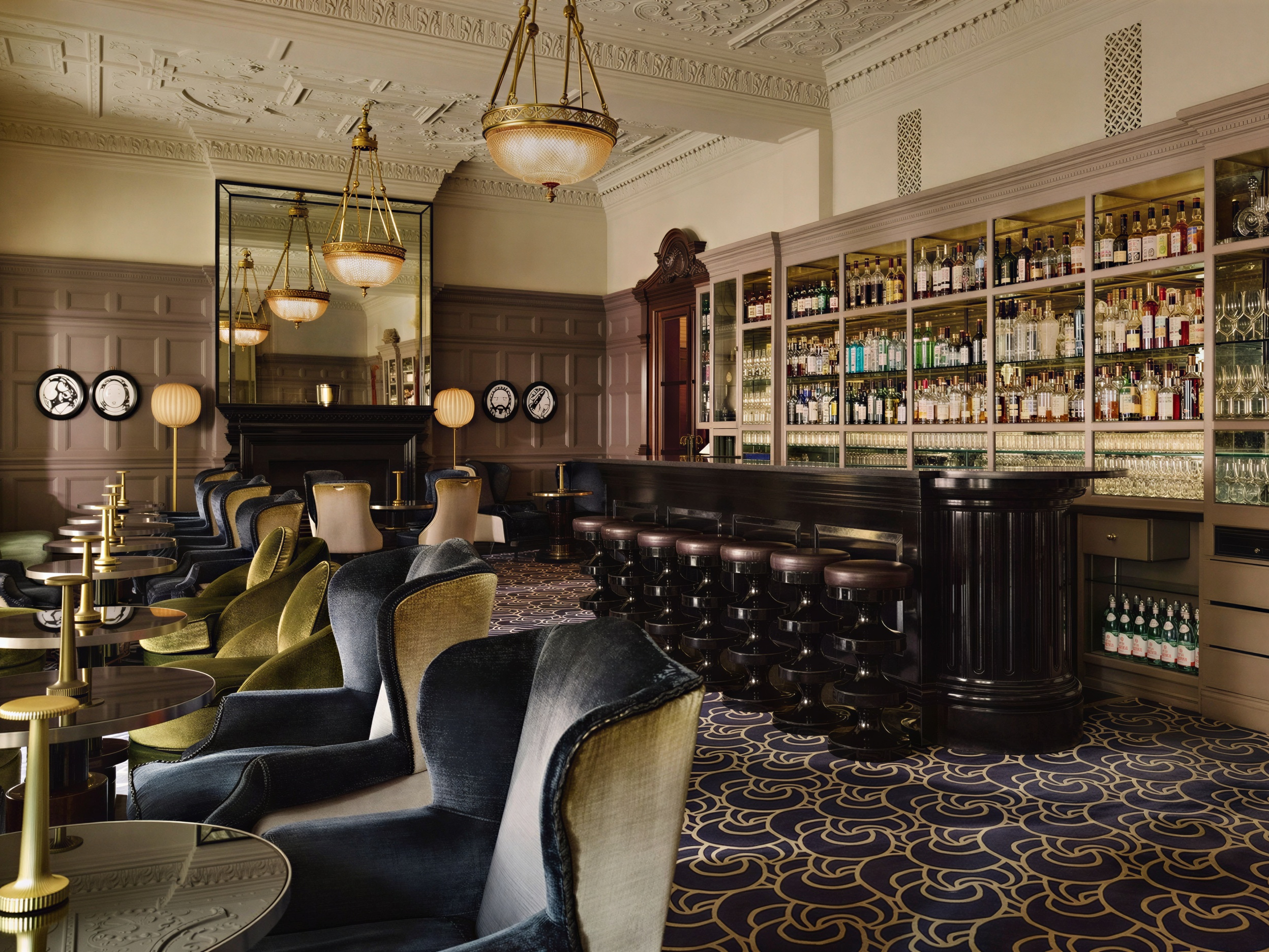 Elegant bar interior with plush armchairs, ornate ceiling, and stocked wooden bar. Mirrors and soft lighting create a cozy, refined atmosphere.