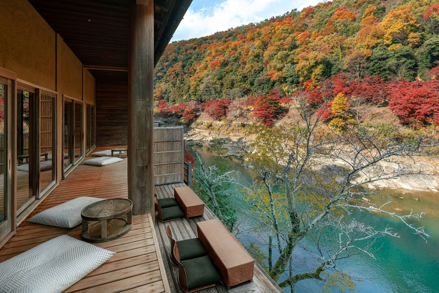 Wooden terrace with cushions and tables, overlooking a river and vibrant autumn foliage on distant hills under a partly cloudy sky.