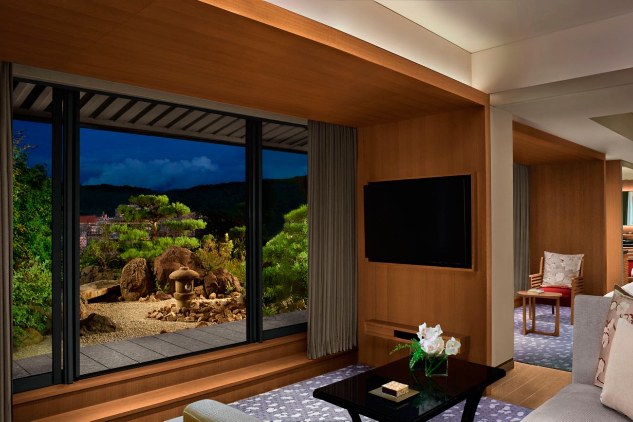 Modern living room with a view of a Japanese garden through large windows. Warm wood tones, a black TV, and floral decor create a serene mood.