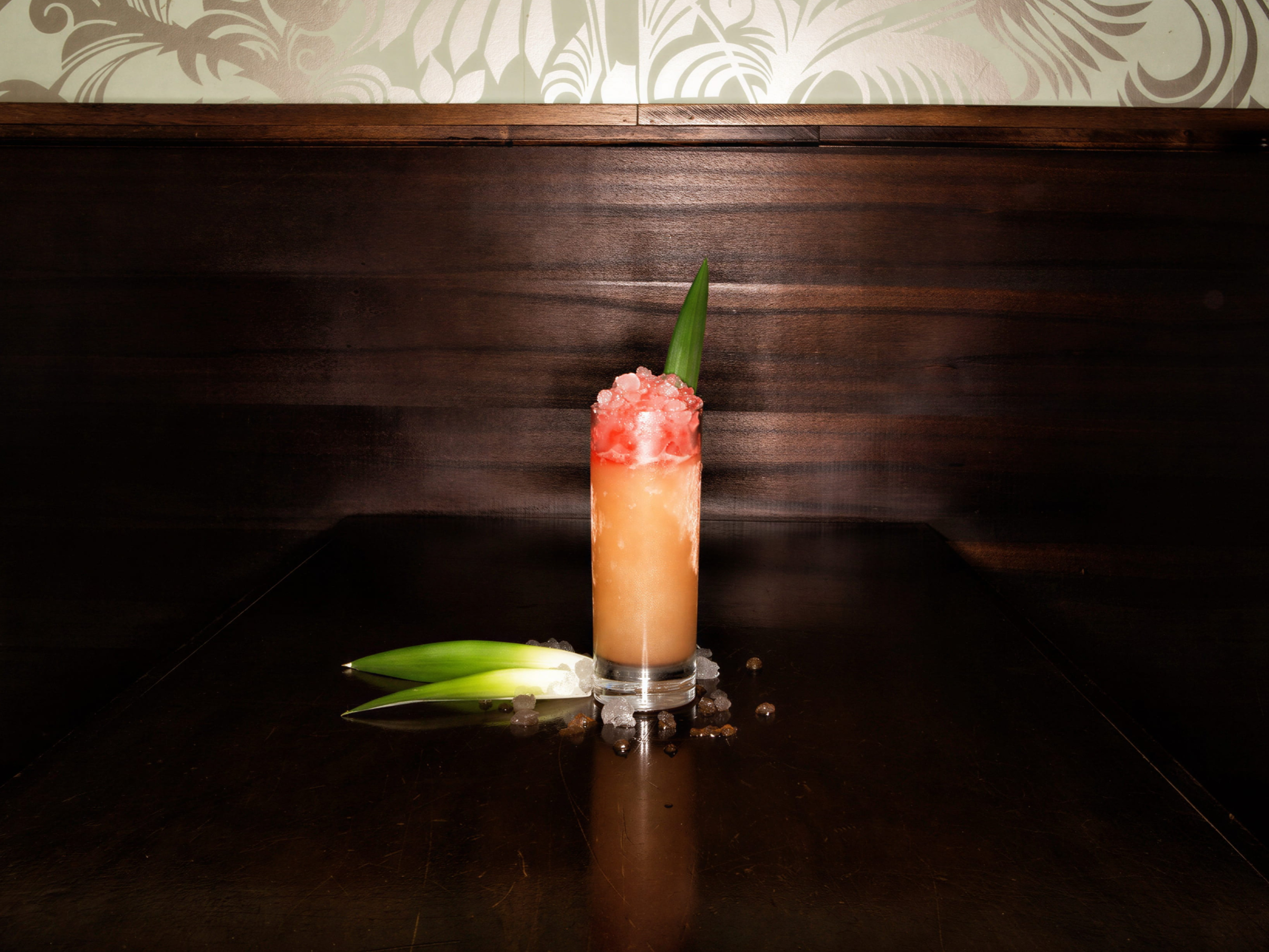 Tall glass with icy pink-orange drink on dark wooden table, green leaf garnish. Wallpaper with floral pattern in background. Elegant setting.