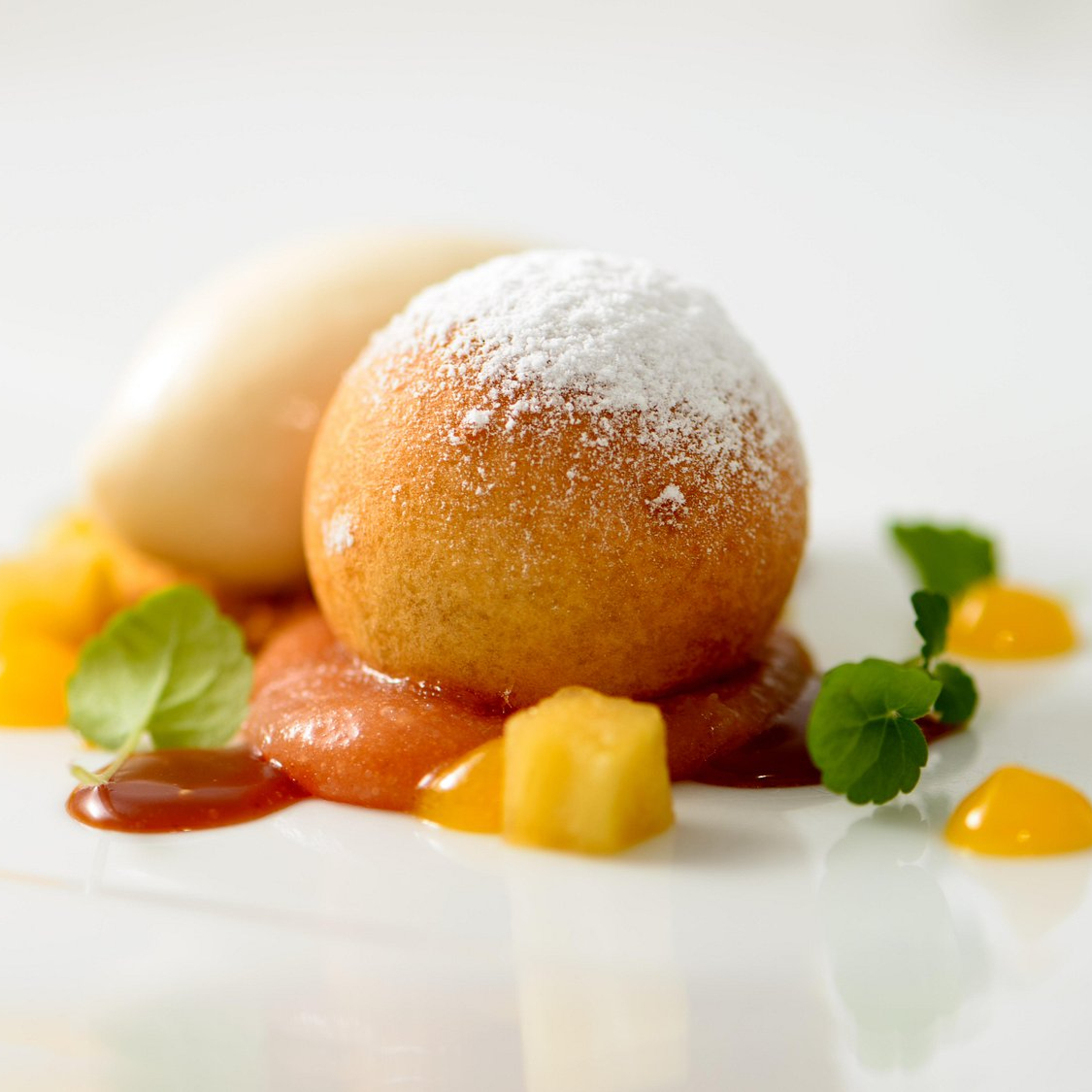 A powdered dessert ball on sauce with fruit cubes and green garnish, set on a white plate, creating a fresh, vibrant mood.