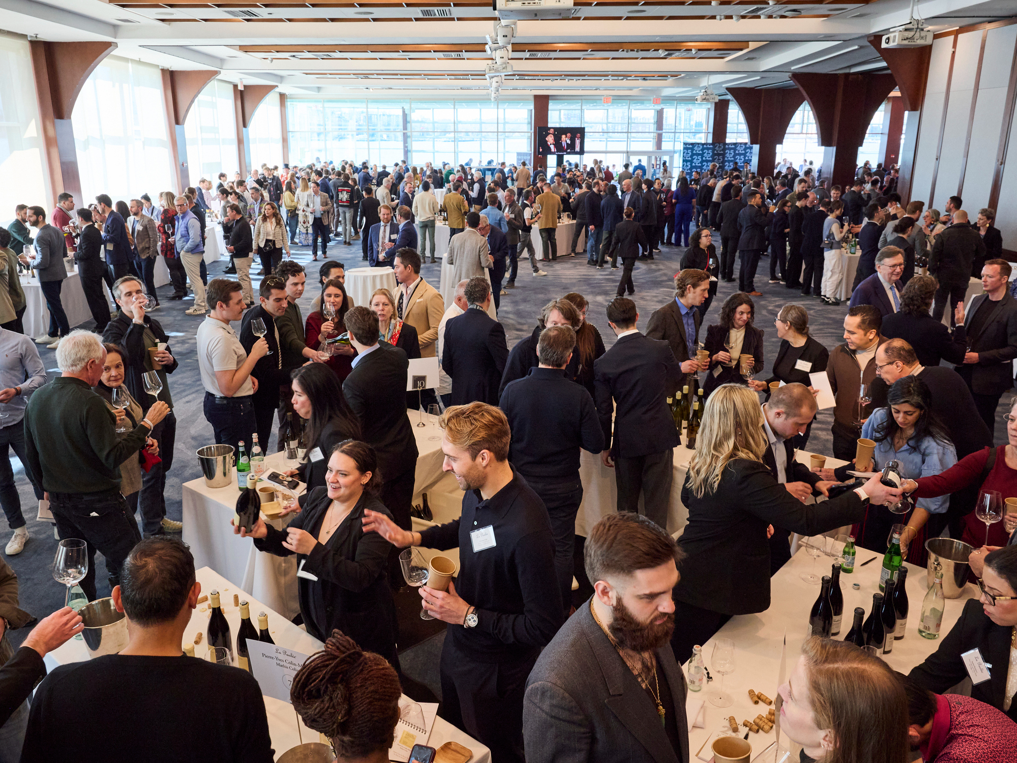 Bright Grand Tasting room at La Paulée NYC 2025. Guests socialize, sampling wine at tables. Attendees wear name badges, creating a lively atmosphere.