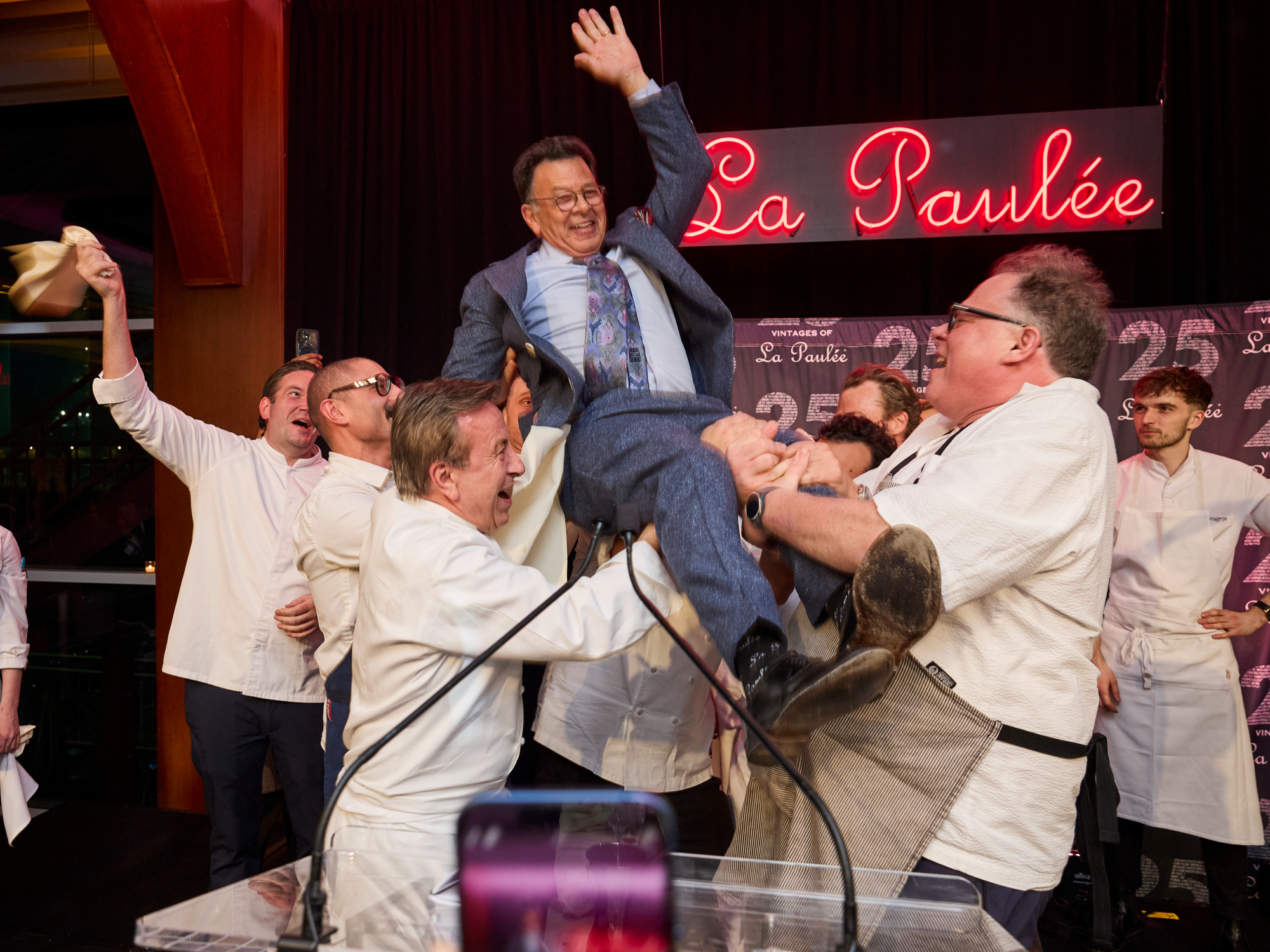 Chefs joyfully lift La Paulée NYC founder Daniel Johnnes n the air at the 2025 Gala Dinner under a neon "La Paulée" sign. Lively atmosphere with a patterned backdrop.