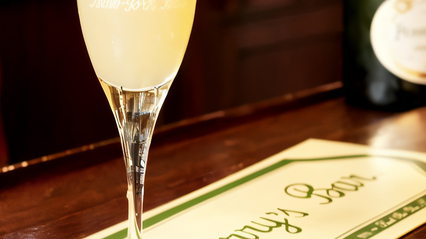 A cocktail in a tall glass sits on a bar mat labeled "Harry's Bar" on a wooden counter. Background features a dark bottle.