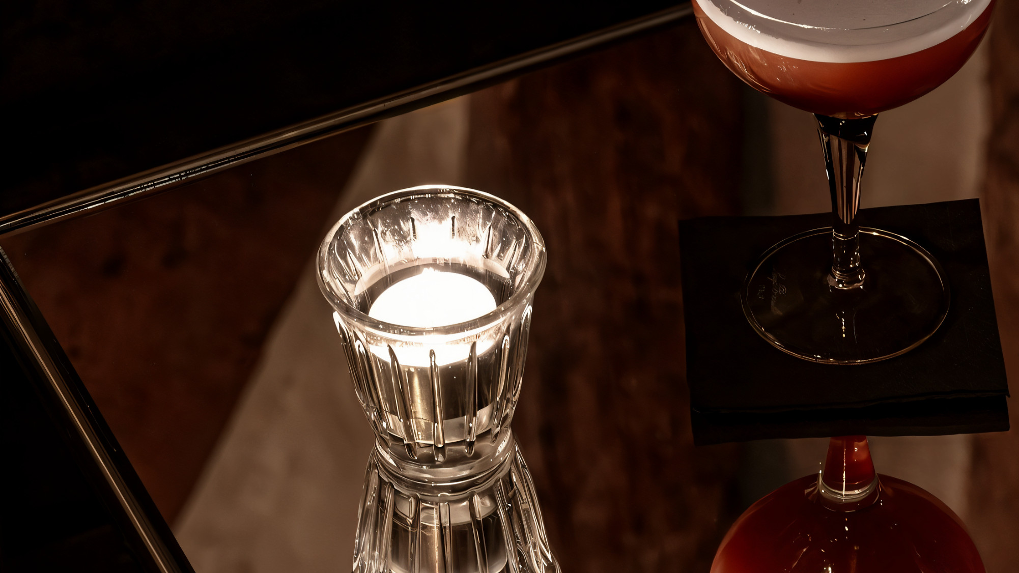 A cocktail glass with a frothy drink and a lit candle are reflected on a glass surface. The scene is dimly lit with a warm, relaxed ambiance.