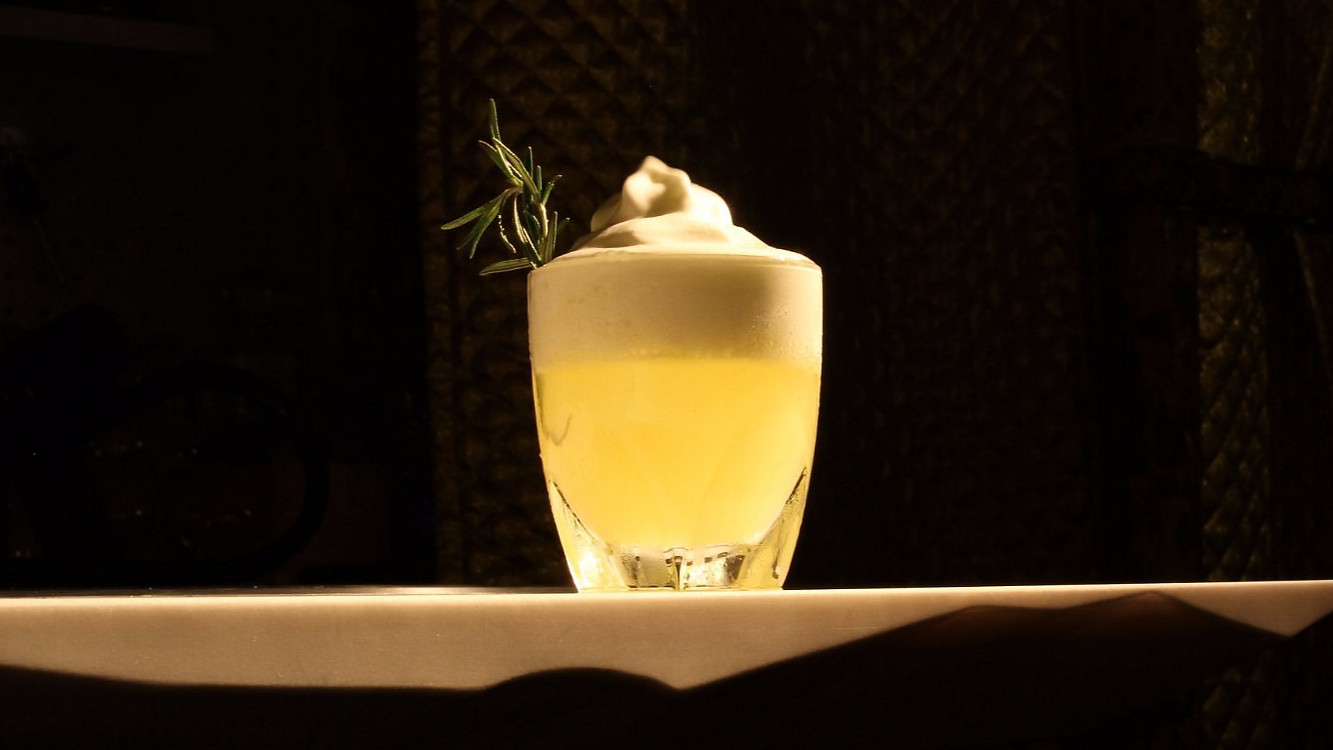 A frothy cocktail garnished with herbs sits on a white marble table beside a crystal decanter. Dim, textured golden background.