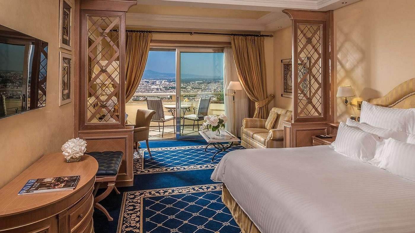 Elegant hotel room with a large bed, blue patterned carpet, and gold curtains. Balcony view shows a cityscape and mountains. Cozy ambiance.
