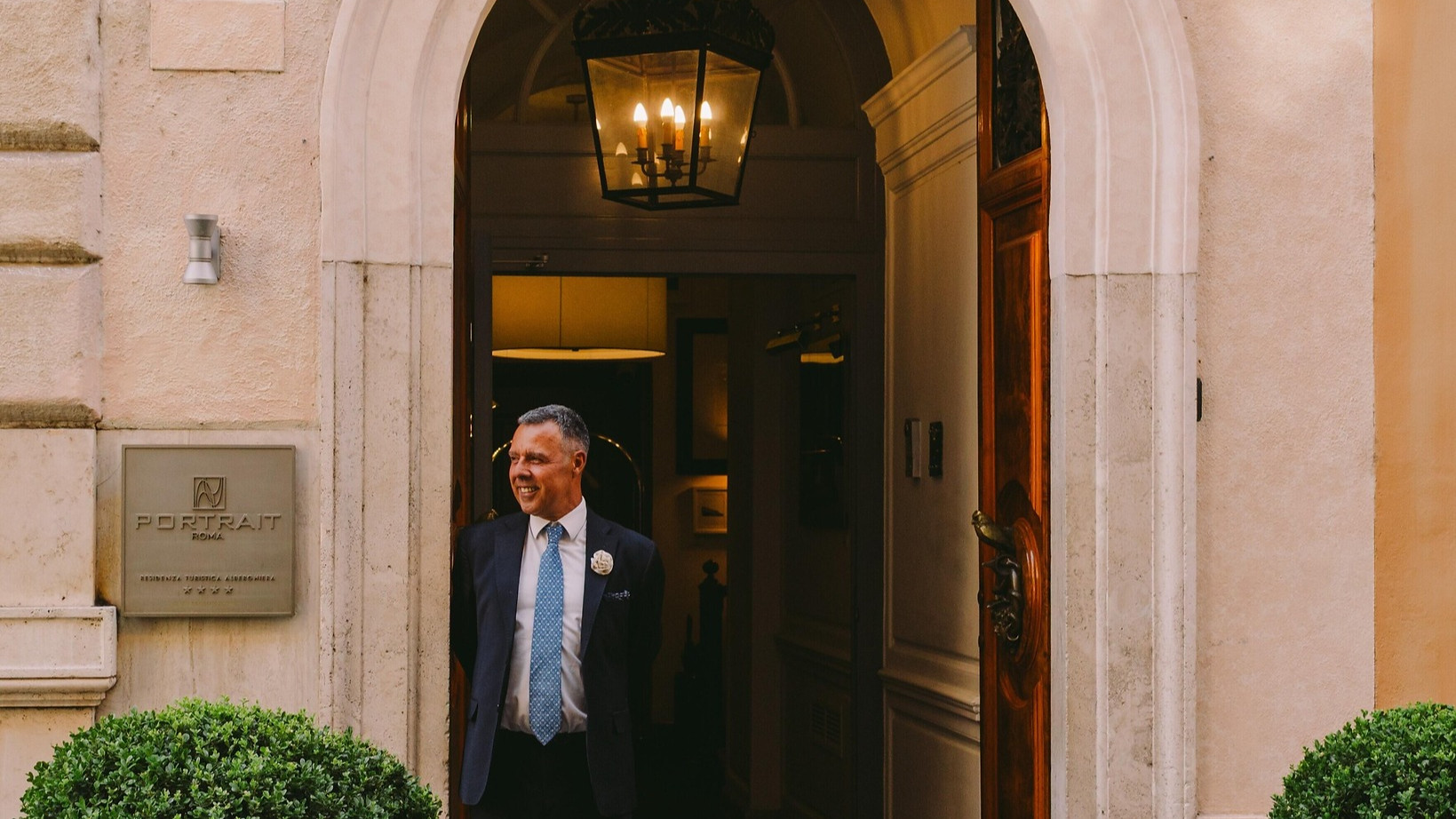 Man in a suit stands smiling under an archway at an entrance labeled "Portrait Roma." Two potted plants flank the doorway, number 23 visible.