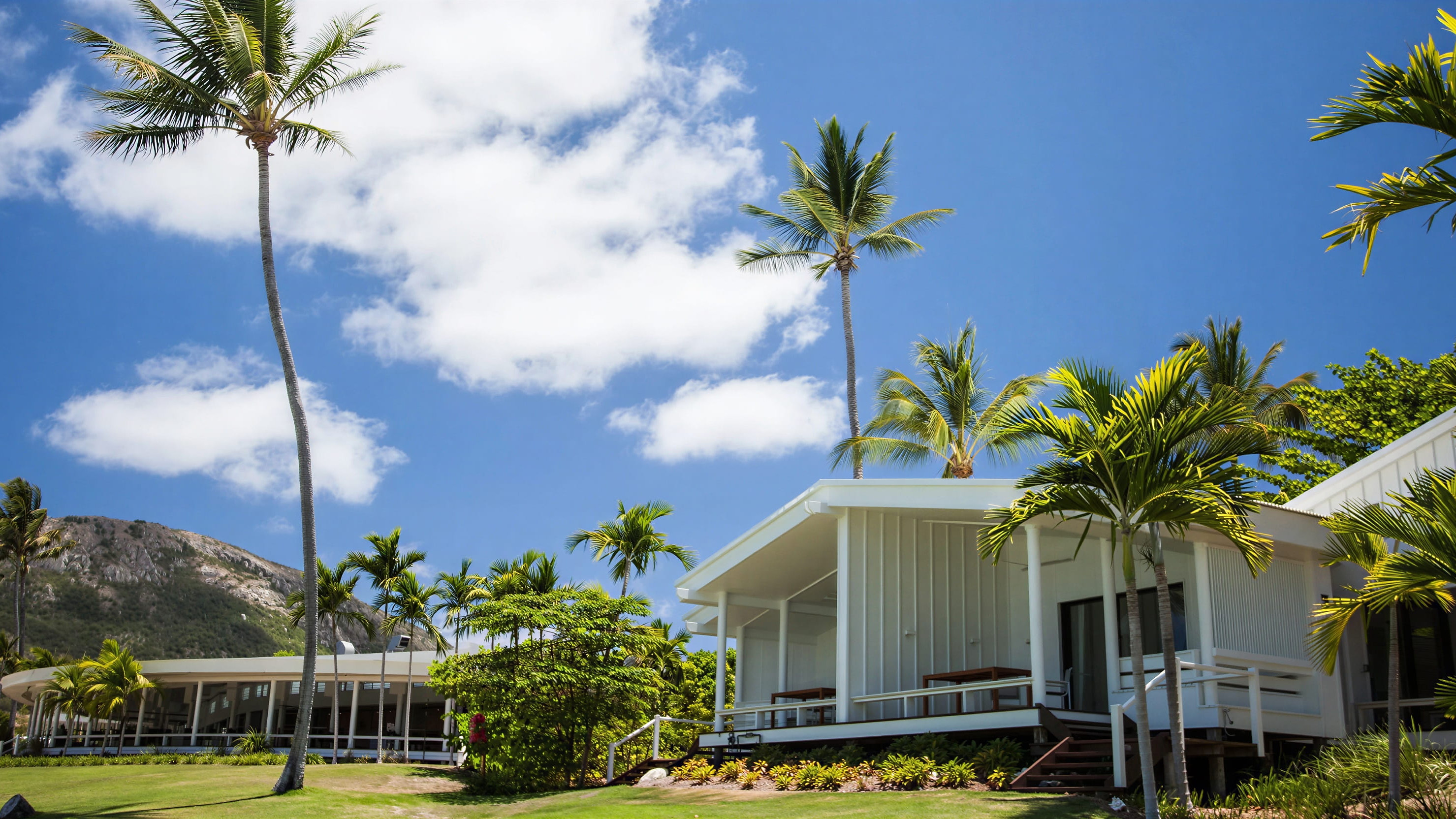 Beachfront Suite exterior at Lizard Island with palm trees, lush greenery, and a mountain backdrop under a bright blue sky with clouds. Peaceful tropical vibe.