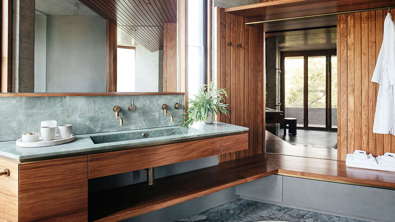 Modern bathroom at The House on Lizard Island Resort with wooden walls, green stone sink, and bronze fixtures. White robes hang neatly. Large window offers natural light. Calm atmosphere.