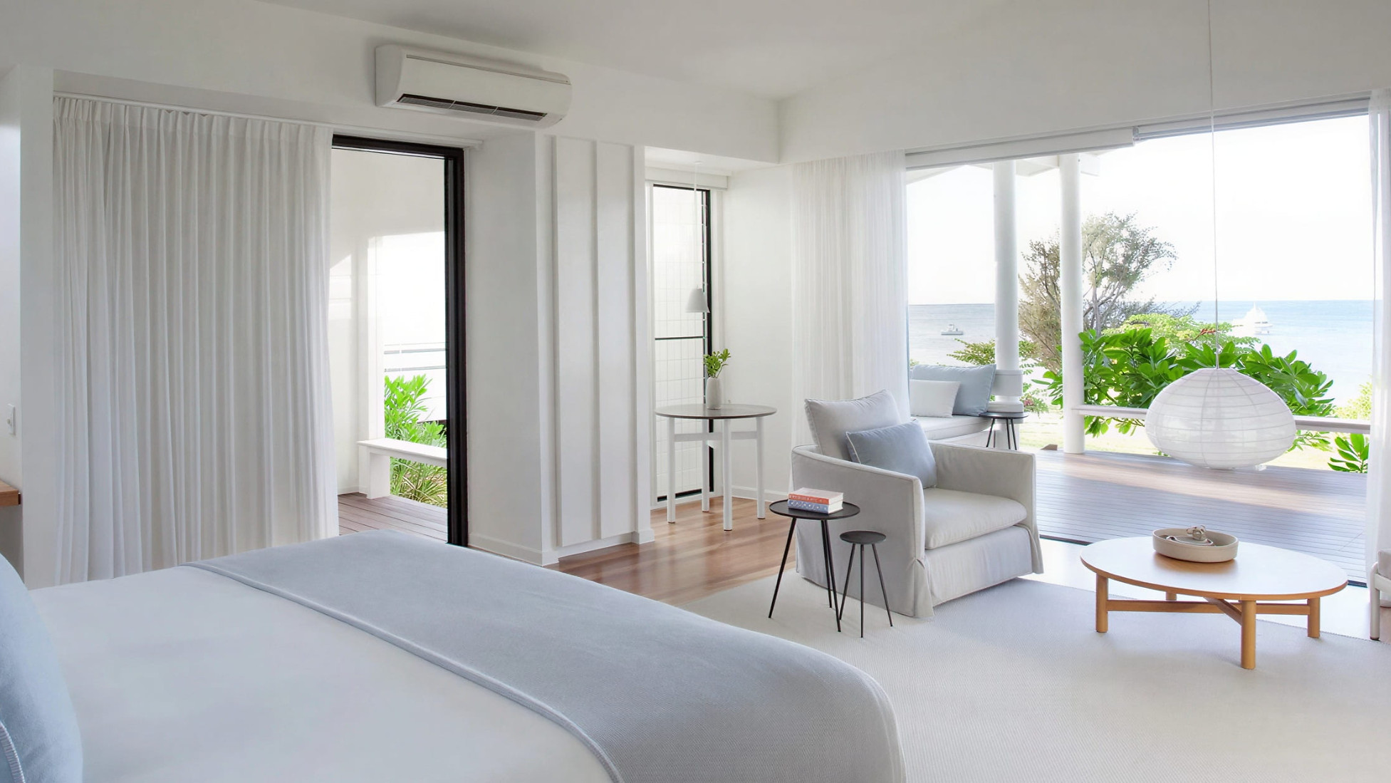 Beachfront Suite interior at Lizard Island Resort with white and light blue decor, ocean view. Features bed, chairs, round table, large windows, and a serene ambiance.