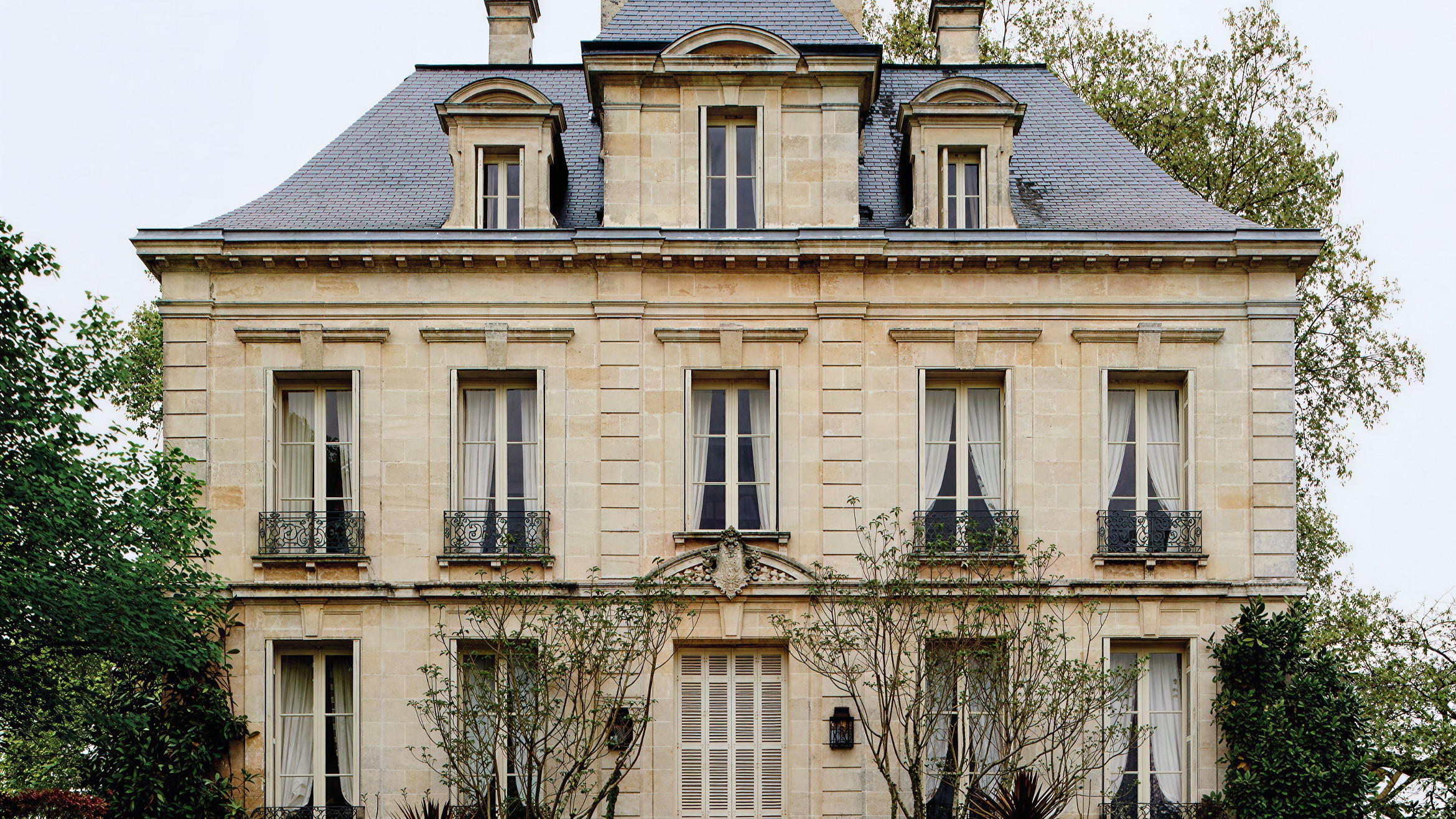 A grand French chateau with stone facade, slate roof, and multiple windows. A manicured garden and steps lead to the entrance.