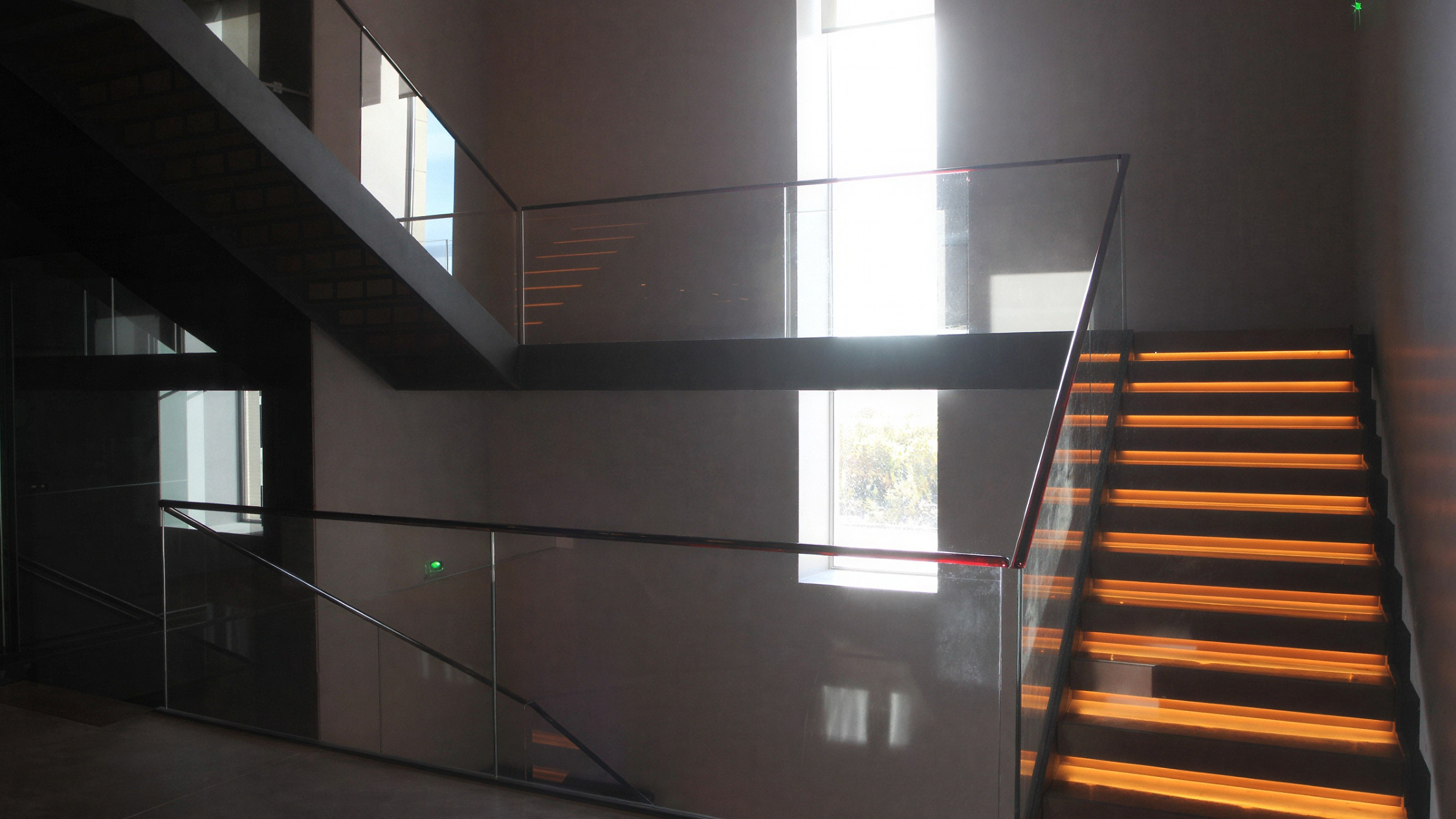 Modern stairwell with glowing orange-lit steps and glass railings, a person climbing stairs, large window, and minimalist design.