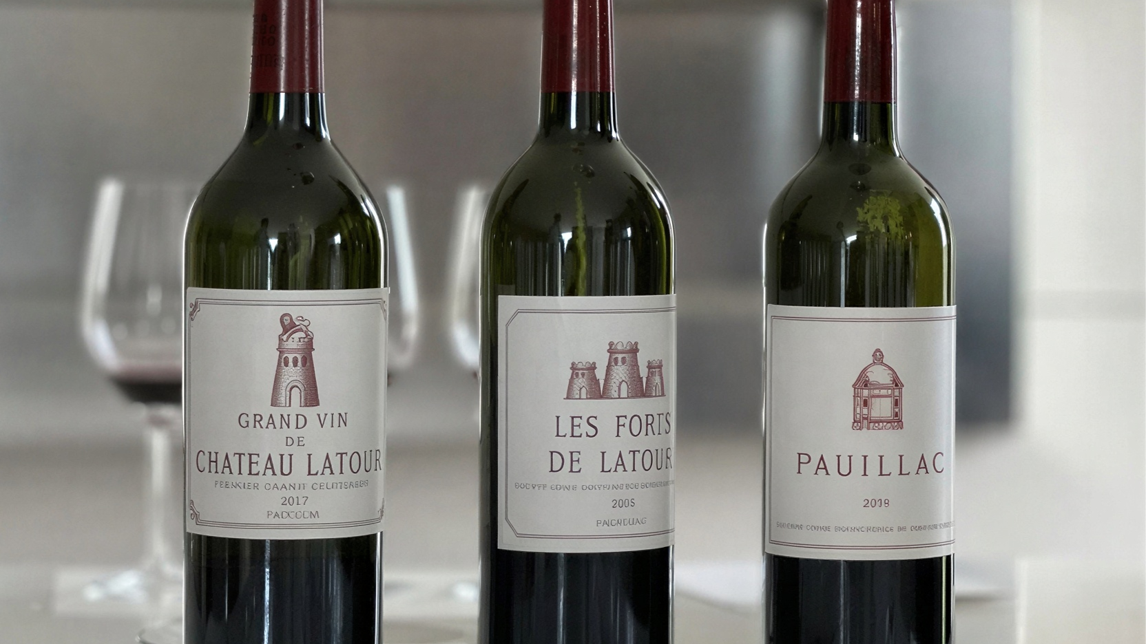 Three wine bottles on a table, labels read: Chateau Latour 2017, Les Forts de Latour 2015, Pauillac 2019. A wine glass and neutral decor in the background.